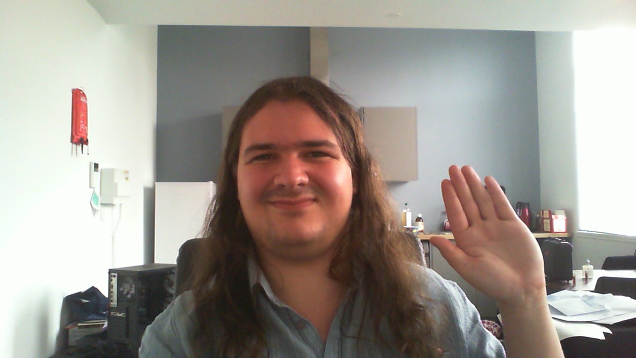 A picture of me, Logan, sat at my desk & waving at the camera.